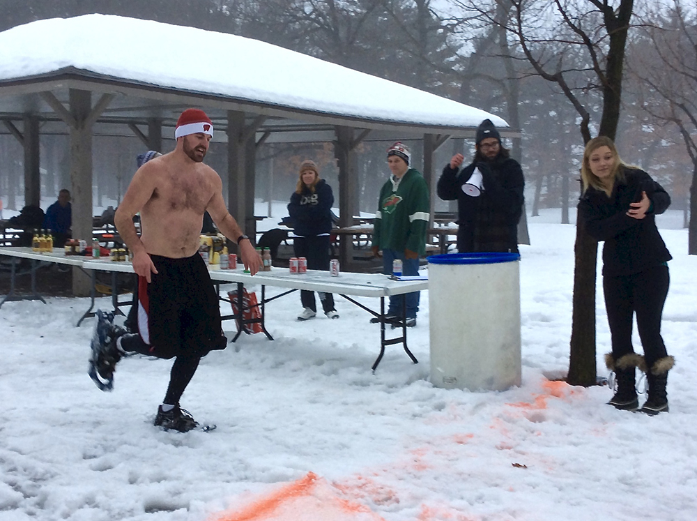 Shirtless man running in snowshoes in snow over a finish line at the Snowshoe Beer Mile.