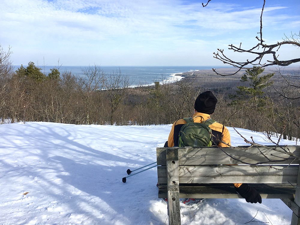 Male snowshoeing in the Porcupine Mountains rests on a bench with an overview of Lake Superior.