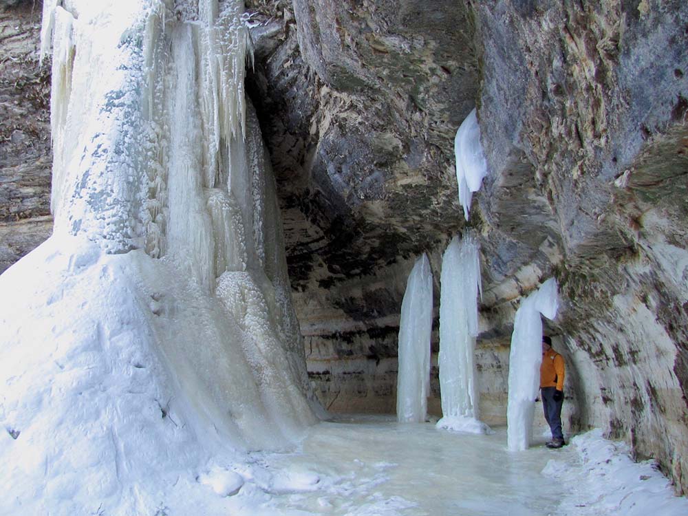 Person standing behind frozen waterfalls pouring over rocks at Pictured Rocks National Lakeshore.