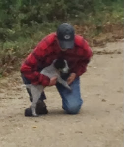 Man in jeans and red-checked shirt kneeling down and holding a puppy on a gravel trail near Kewaunee.