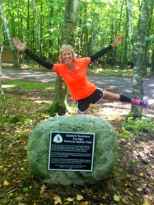 Woman in an orange shirt and black shorts making the star pose near a rock with a plaque stating this is the eastern terminus of the Ice Age Trail.