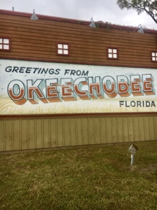 Signing on the side of a red brick building reading, "Greeting from Okeechobee Florida." Near Platts Bluff