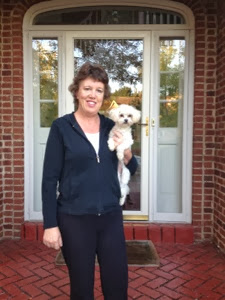 Woman holding a small, white poodle in front of a red-brick home with a white front door.