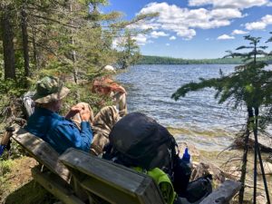 Backpacker sitting on a chair, and another on a rock, at the edge of Loon Lake in the Boundary Waters Canoe Area Wilderness.