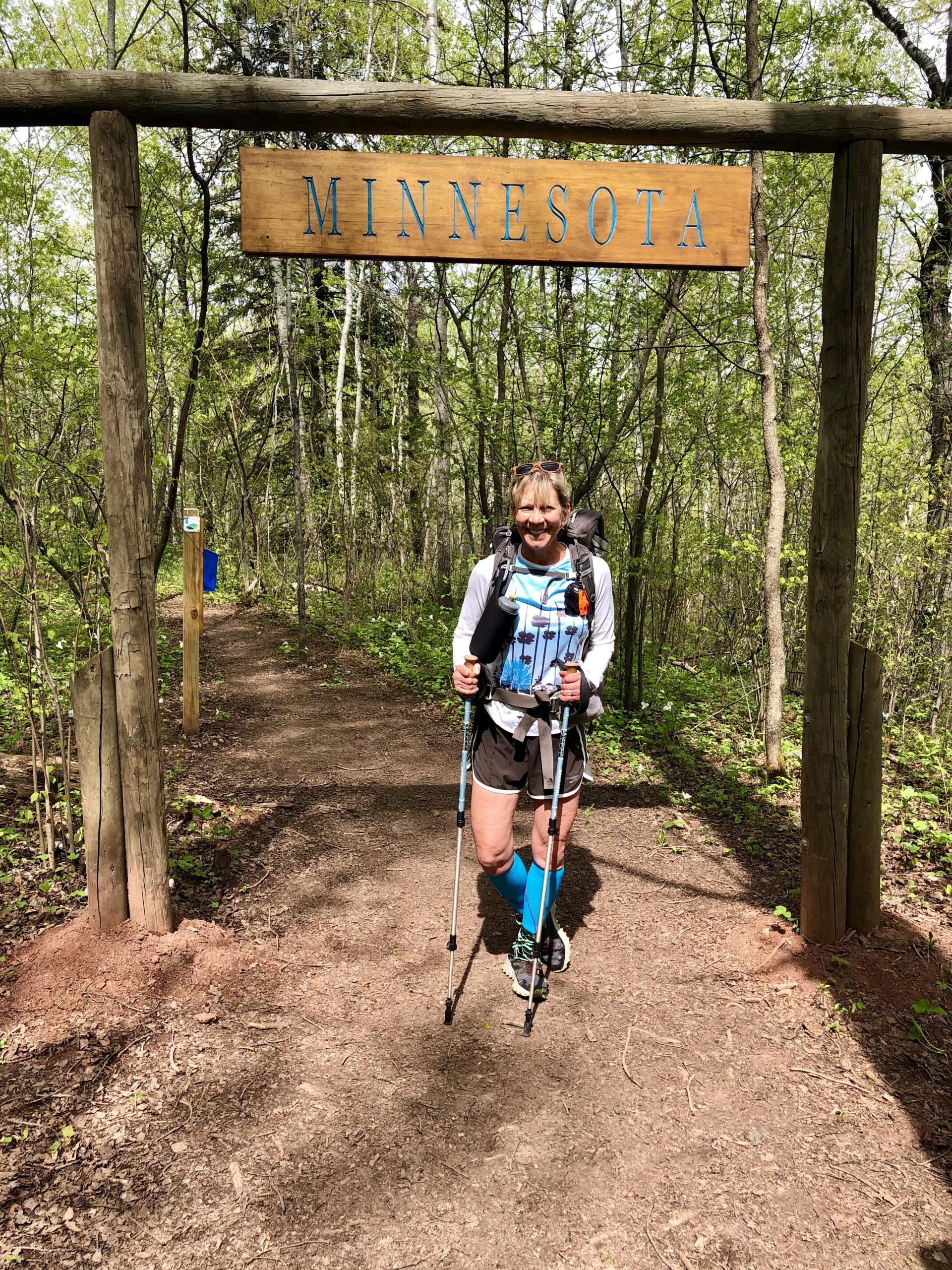 Female hiker on the North Country Trail standing under a sign in the woods saying, "Minnesota."