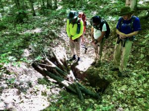 Three hikers on North Country Trail near Valley Spur, Mich., stand by a rectangular hole in the ground with logs over it.
