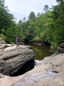 Woman standing on huge rock near waterfalls and green trees en route to Black River Harbor.