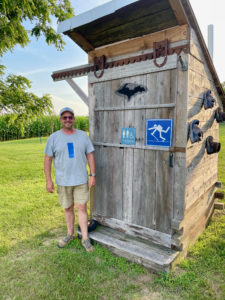 Man standing in front of a wooden outhouse with hiking signs and shoes decorating it.