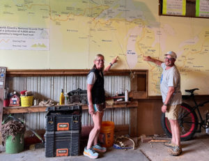 Two people pointing to a large map of the North Country Trail, mounted on a garage wall.