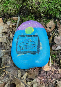 Painted rock on forest floor that says, "Be happy."