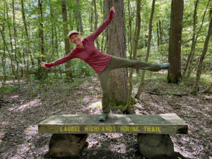 Woman standing on one leg with her arms outstretched on top of a bench in the woods.
